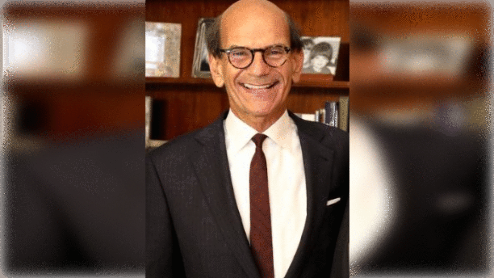 Smiling man in a suit with glasses stands in front of a bookshelf, showcasing a friendly demeanor and professional appearance.