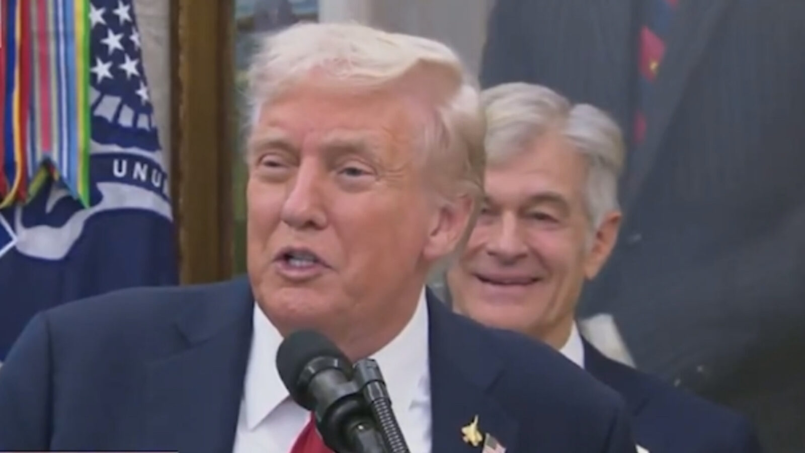 Donald Trump speaking at a podium during an event, with a smiling individual in the background, surrounded by official decorations and flags.