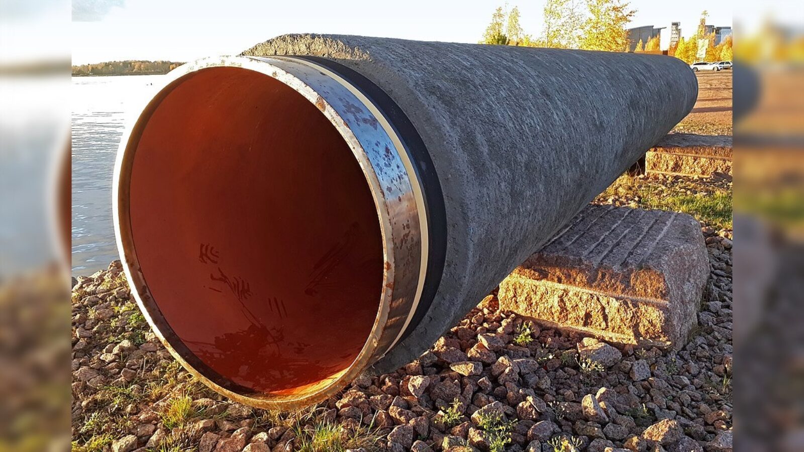 Large concrete pipe resting on a stone base near a body of water, showcasing a rusty interior and surrounded by pebbles and grass.