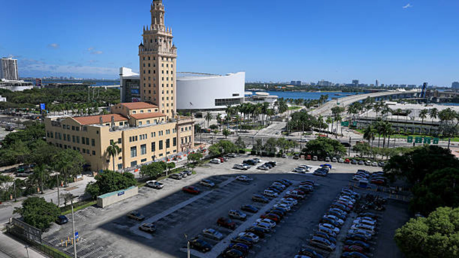 Aerial view of Miami's Freedom Tower alongside a parking lot, with modern buildings and palm trees in the background under a clear blue sky.
