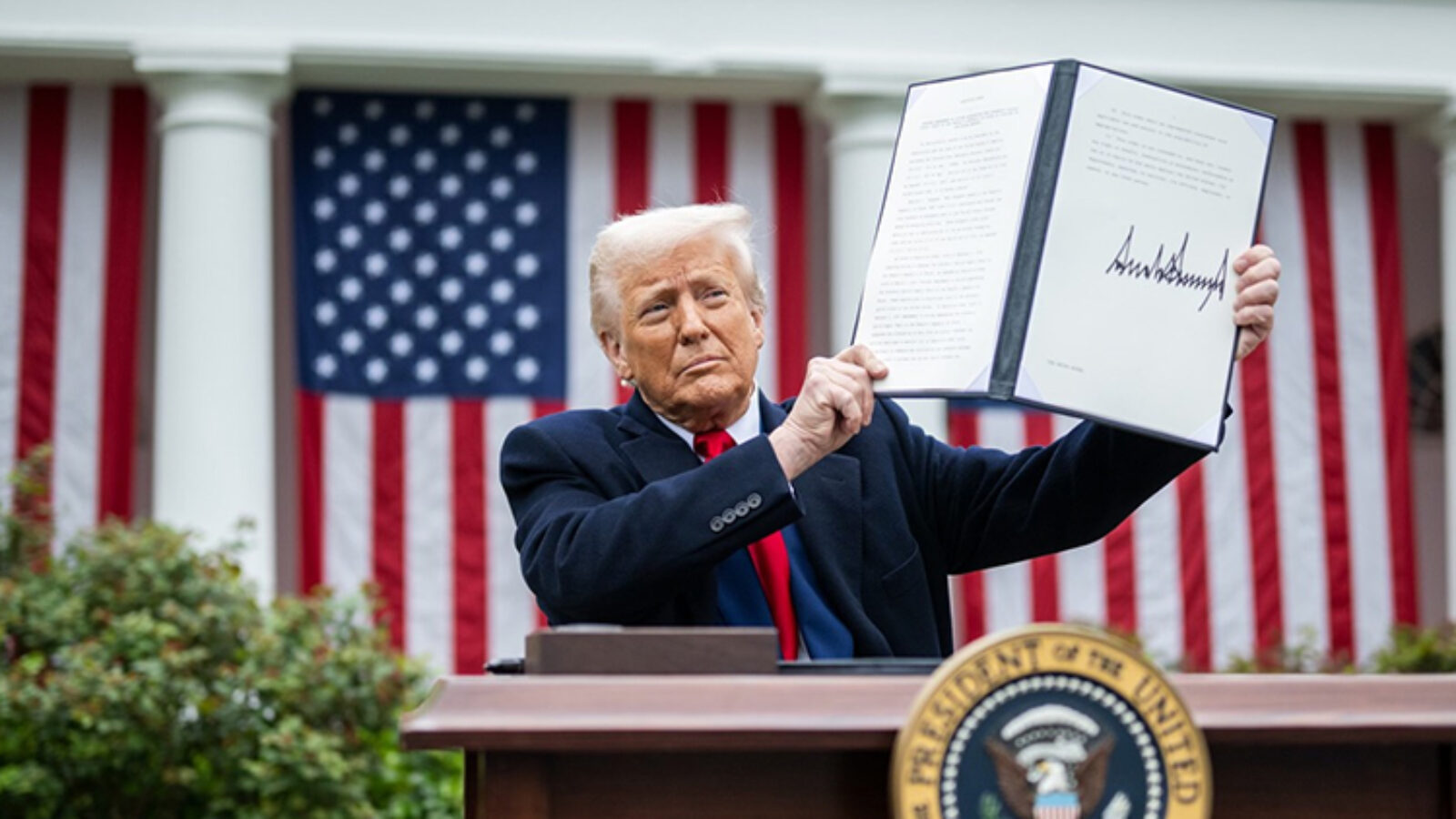 Donald Trump holds up a signed document at a podium in front of American flags, showcasing a significant announcement.