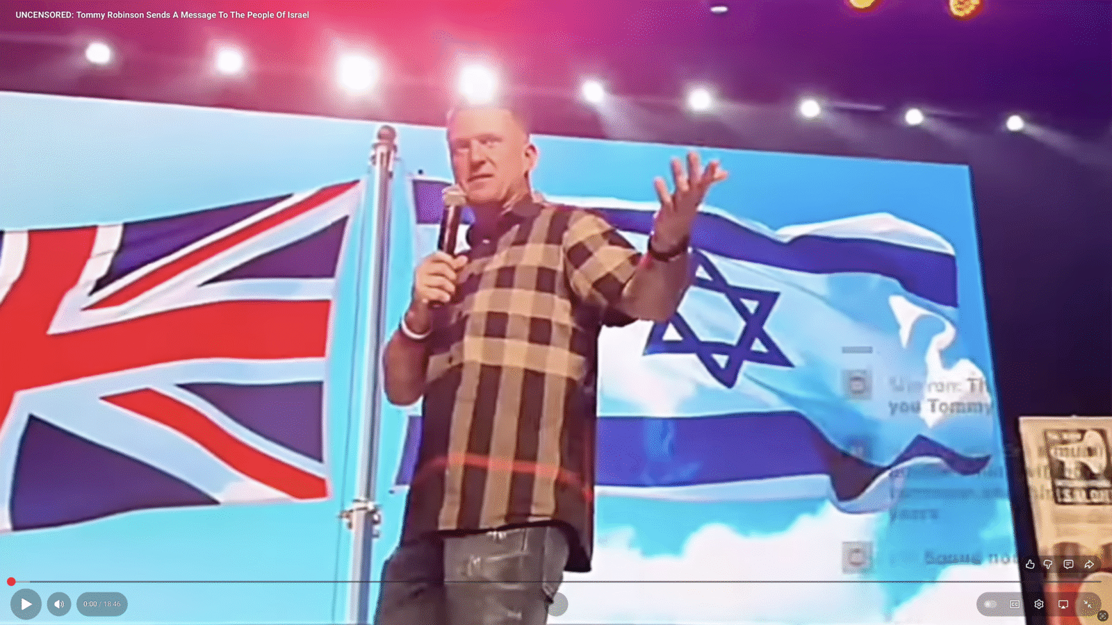 Tommy Robinson speaking on stage with British and Israeli flags in the background, addressing an audience at a public event.