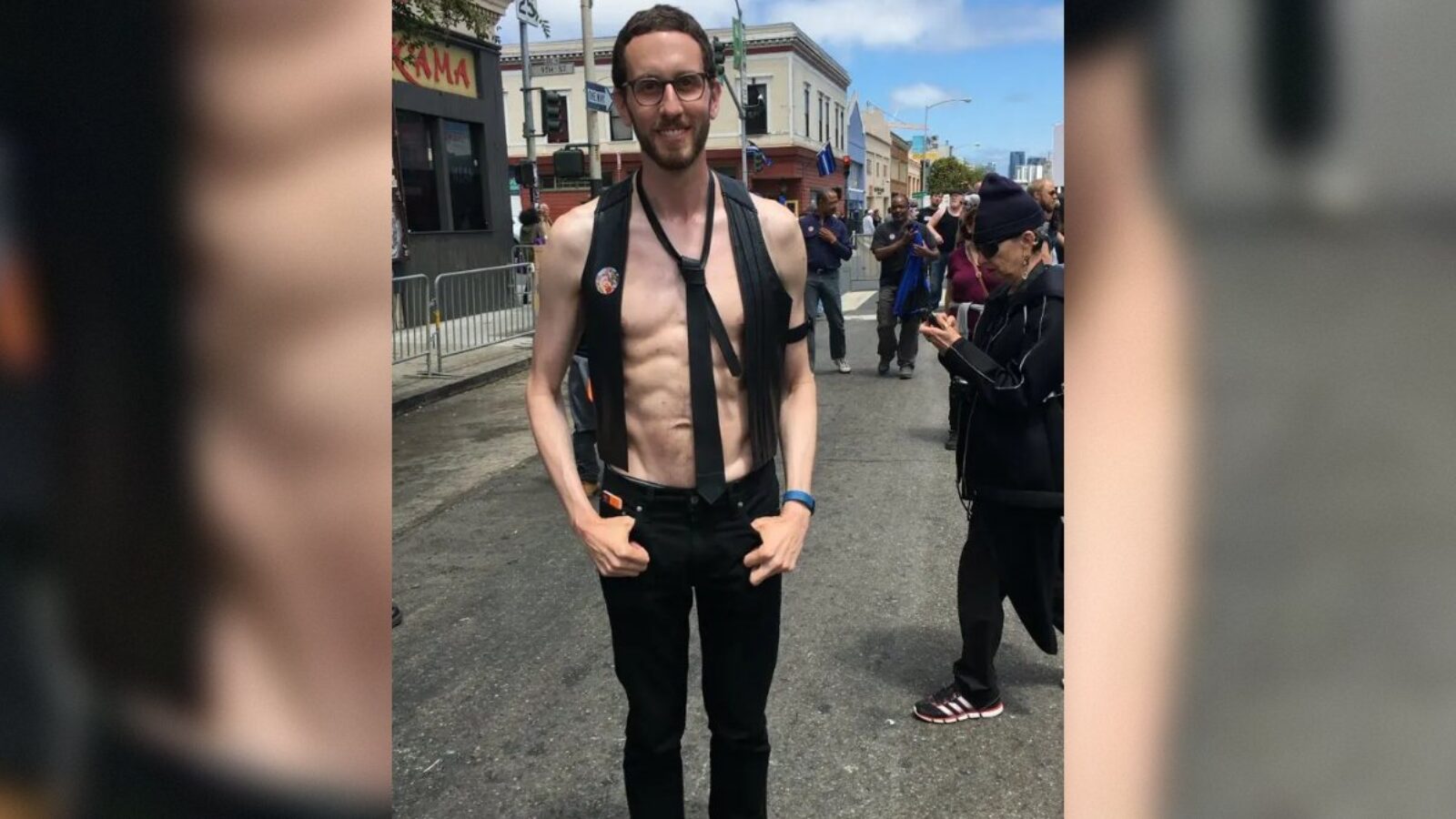 Man wearing a harness and black pants stands confidently on a city street during a lively outdoor event, surrounded by onlookers.