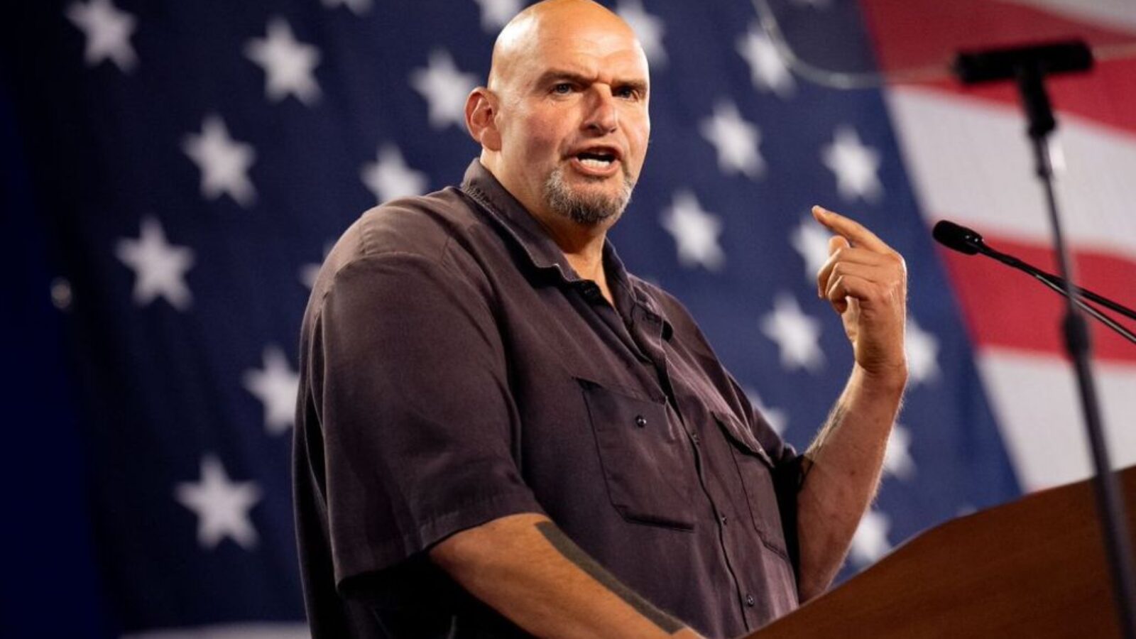 Man speaking passionately at a podium with an American flag backdrop, emphasizing key points during a public address.