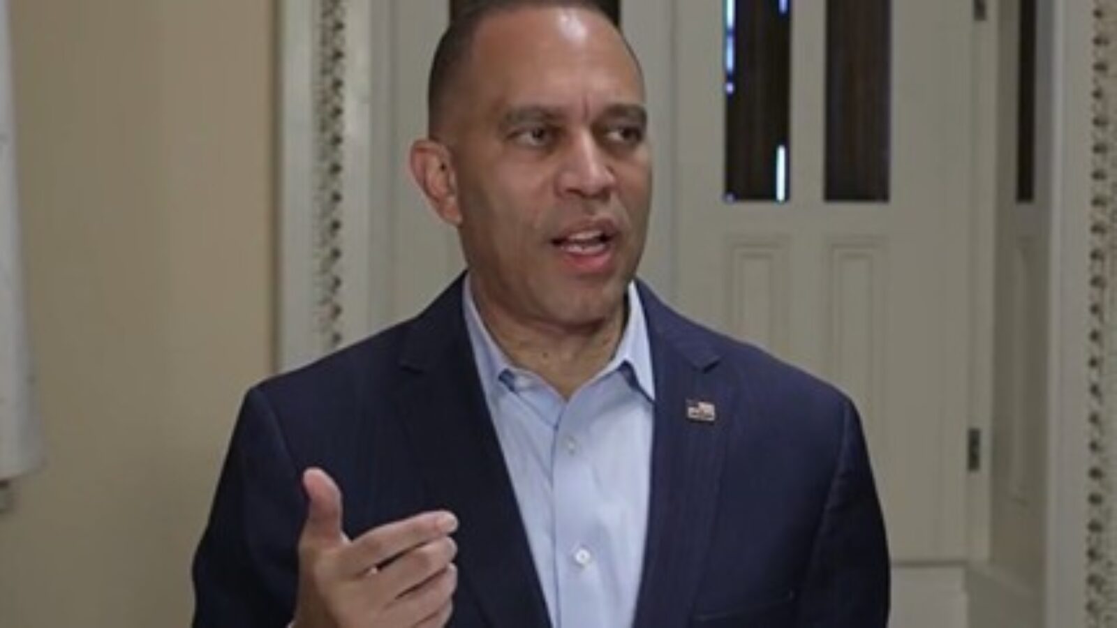 Man in a blue suit gestures while speaking in a well-lit indoor setting, emphasizing key points during a discussion.