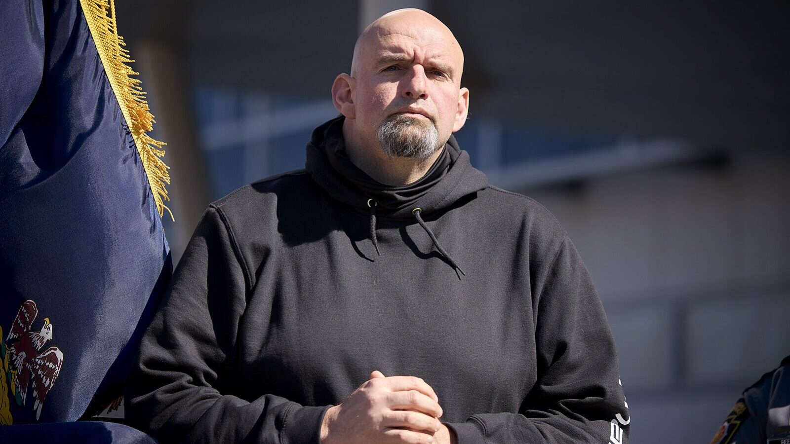 Man in a black hoodie stands confidently with hands clasped, framed by a flag, during an outdoor event in bright sunlight.