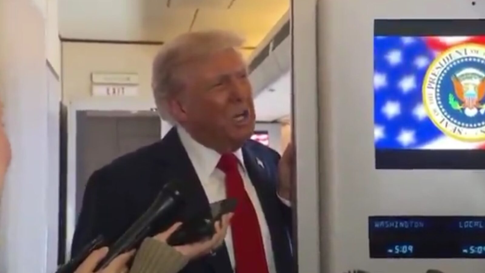 Donald Trump speaking to reporters on an airplane, with a presidential seal displayed on a screen in the background.