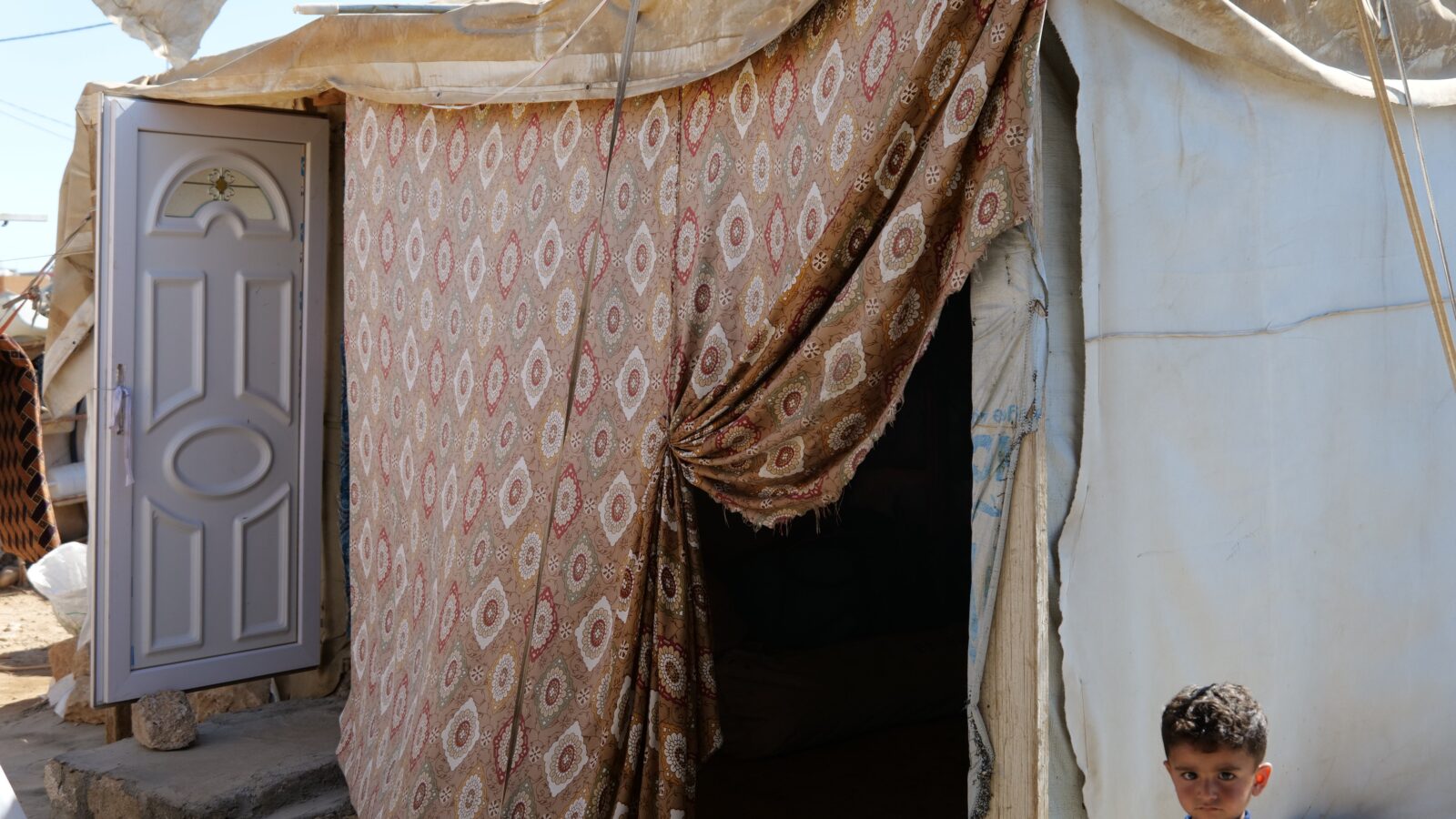 A young boy stands outside a patterned tent with an open door, highlighting living conditions in a refugee camp.