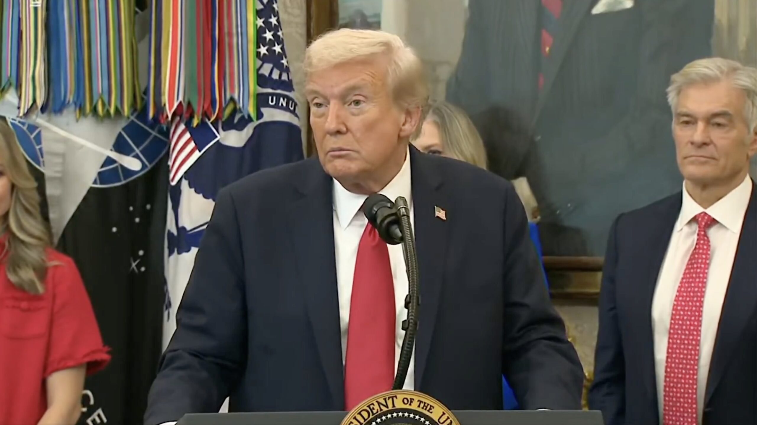 Donald Trump speaks at a podium in front of military decorations, flanked by supporters during a press event.