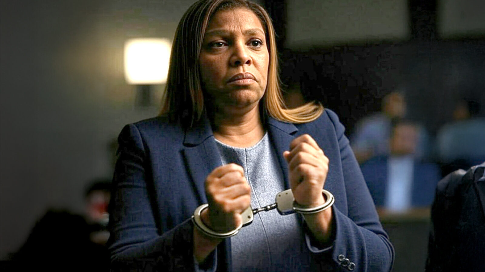 A woman in handcuffs, wearing a navy blazer, appears serious and focused in a courtroom setting.
