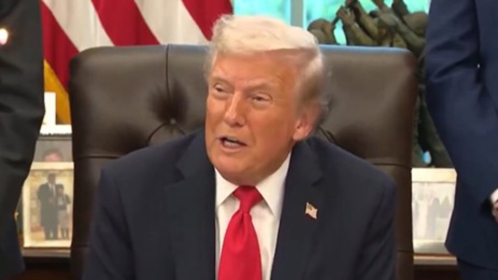 Donald Trump seated in the Oval Office, wearing a suit and red tie, with American flags and historical artifacts in the background.