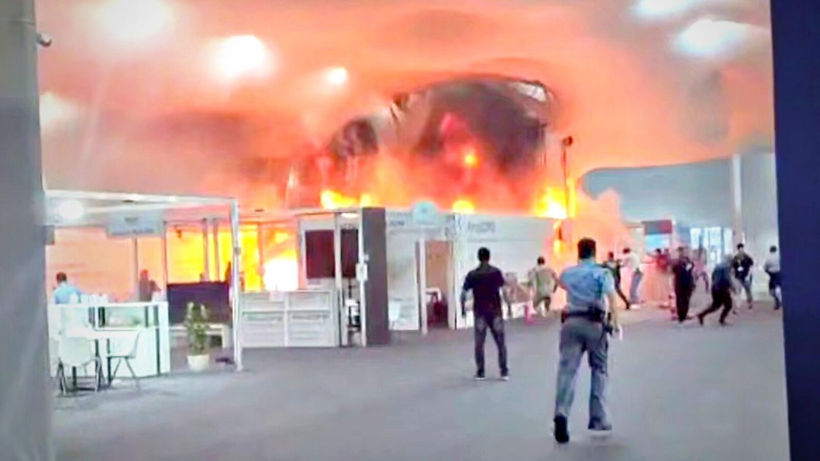 Fire engulfs an exhibition hall as attendees evacuate, showcasing a chaotic emergency situation with thick smoke and flames.