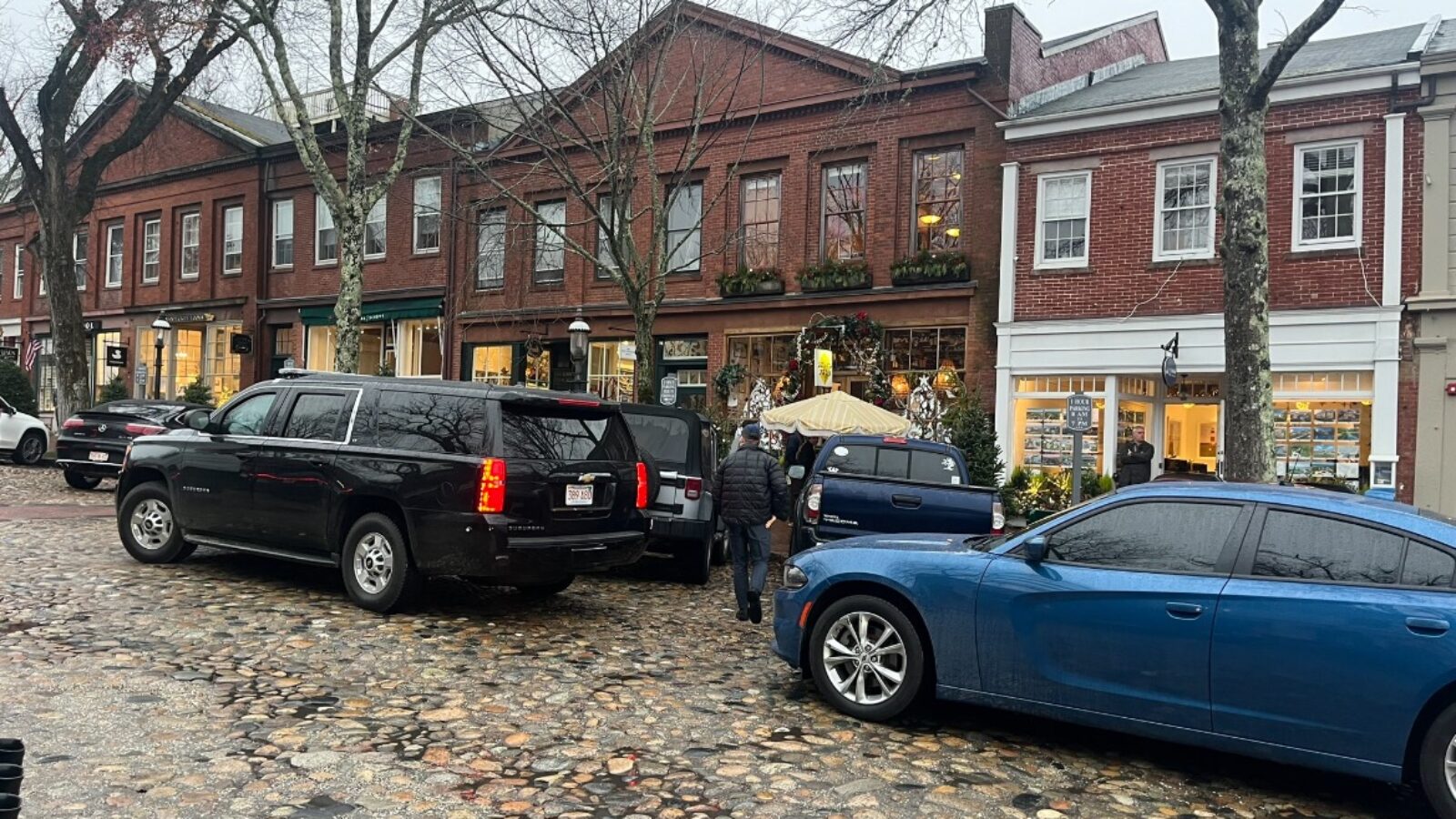 Cobblestone street scene featuring parked vehicles and charming brick storefronts adorned with holiday decorations on a rainy day.