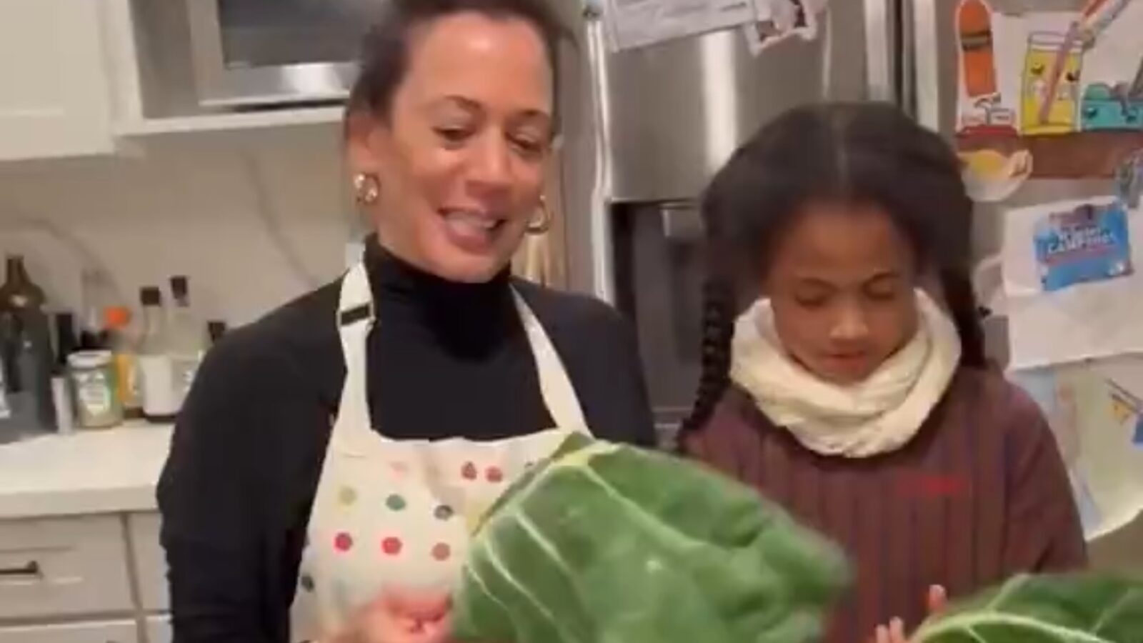 A woman and a girl smile while holding large collard green leaves in a cozy kitchen setting.