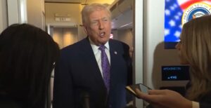 Donald Trump speaking to reporters on an airplane, with a presidential seal visible in the background.