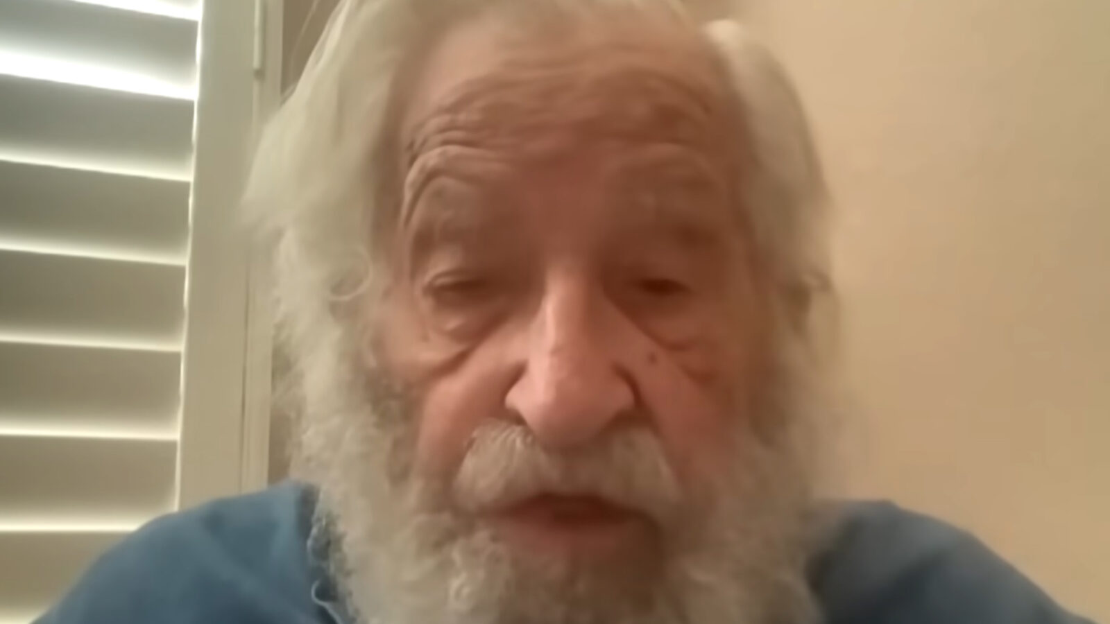 Elderly man with white hair and beard speaking in a video call, showcasing a thoughtful expression against a light background with closed shutters.