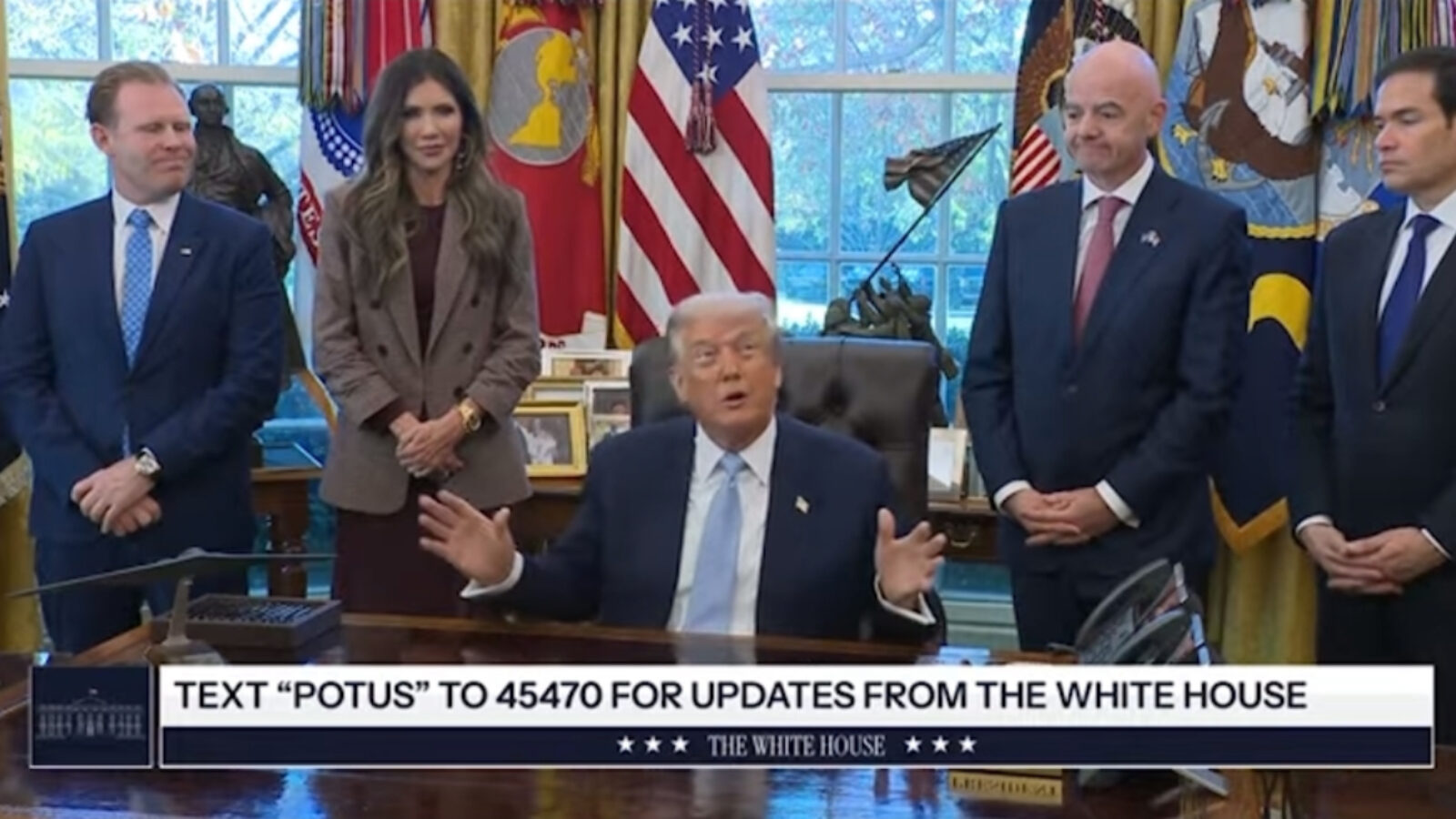 Former President Donald Trump speaks in the Oval Office surrounded by advisors and flags, promoting text updates from the White House.