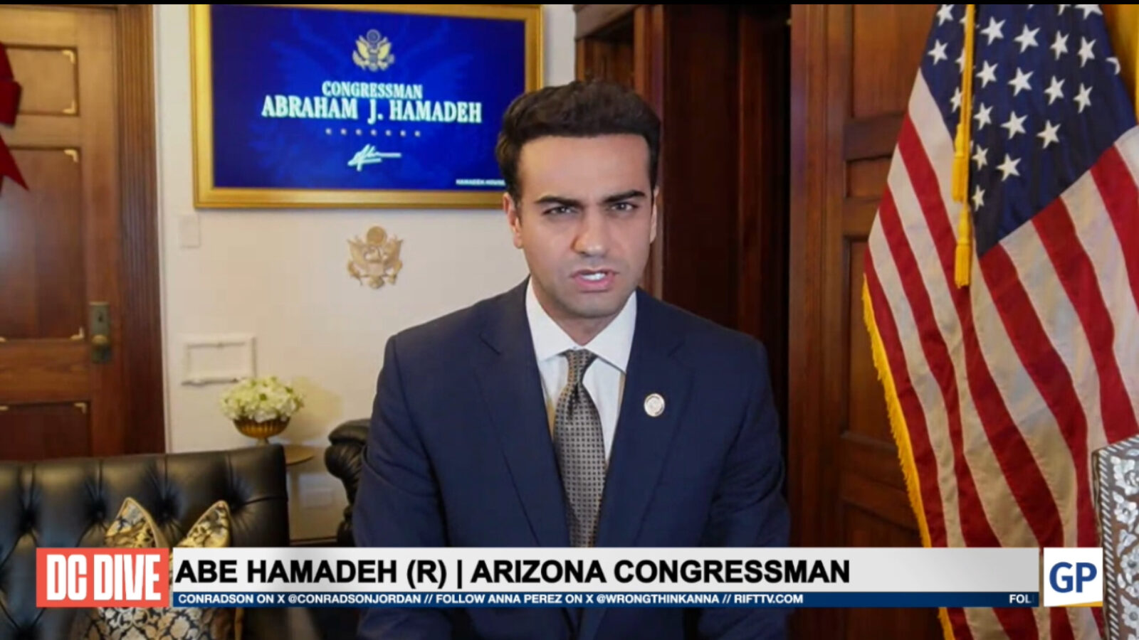 Abe Hamadeh, Arizona Congressman, speaks during a broadcast from his office, featuring an American flag and his nameplate in the background.