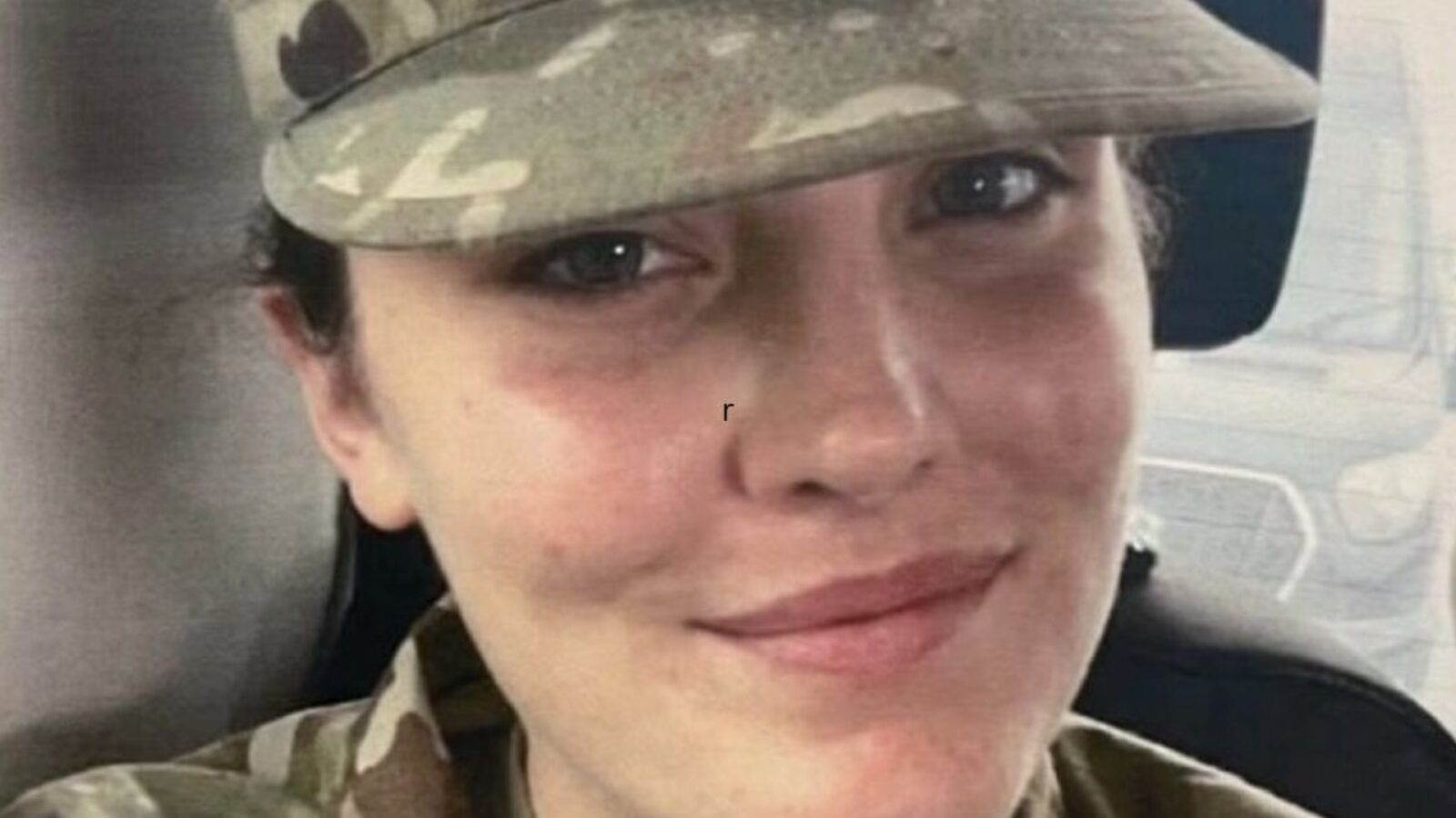 Smiling military personnel wearing a camouflage cap, seated inside a vehicle, showcasing a friendly demeanor.