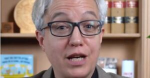 Person speaking directly to the camera with light-colored hair and glasses, set against a background of books and decorative items in a well-lit room.