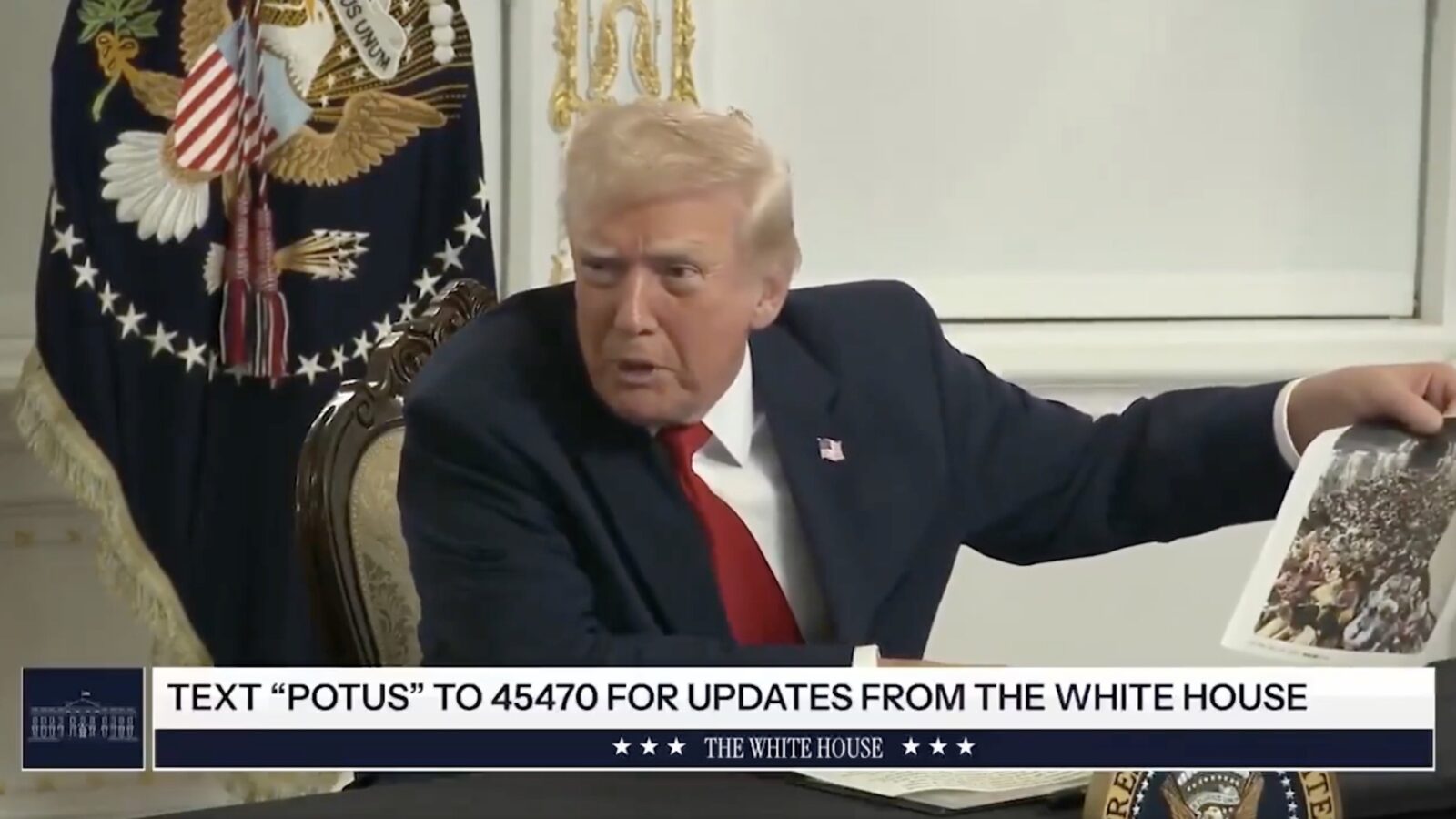 Donald Trump gestures while discussing a photo during a meeting at the White House, featuring the presidential seal in the background.
