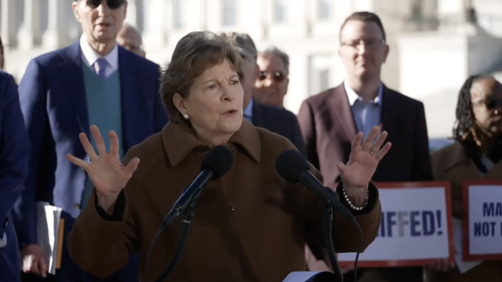 A woman speaks passionately at a press conference, gesturing with her hands, while supporters hold signs in the background.