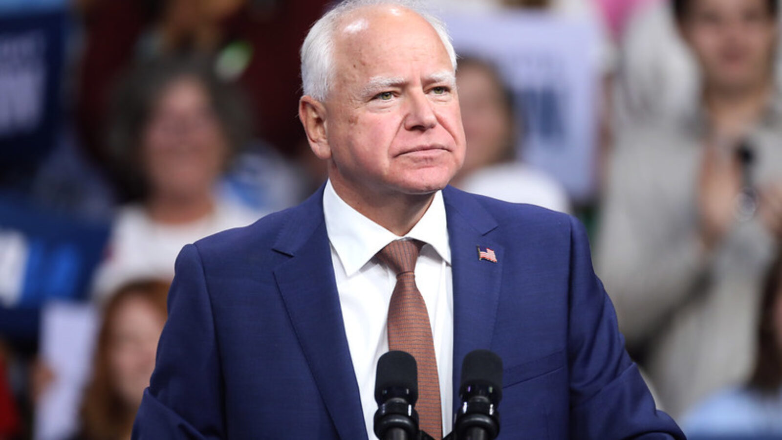 Minnesota Governor Tim Walz speaks at a rally, addressing an engaged crowd with campaign signs in the background.