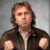 Man with tousled hair and a serious expression, wearing a brown jacket, poses with clenched fists against a textured gray background.