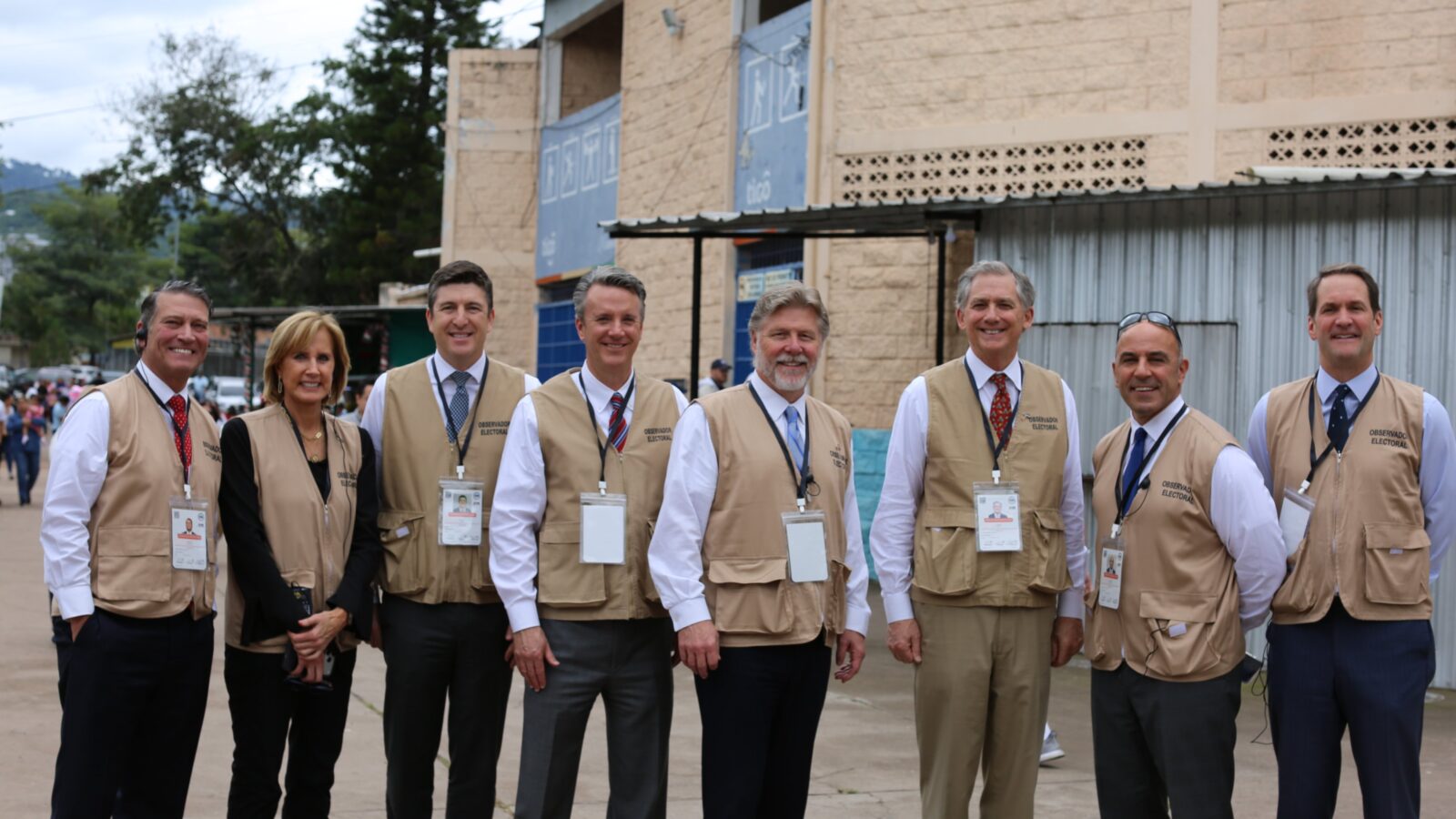 Group of election observers in beige vests standing outdoors, smiling for the camera with a backdrop of a community setting.