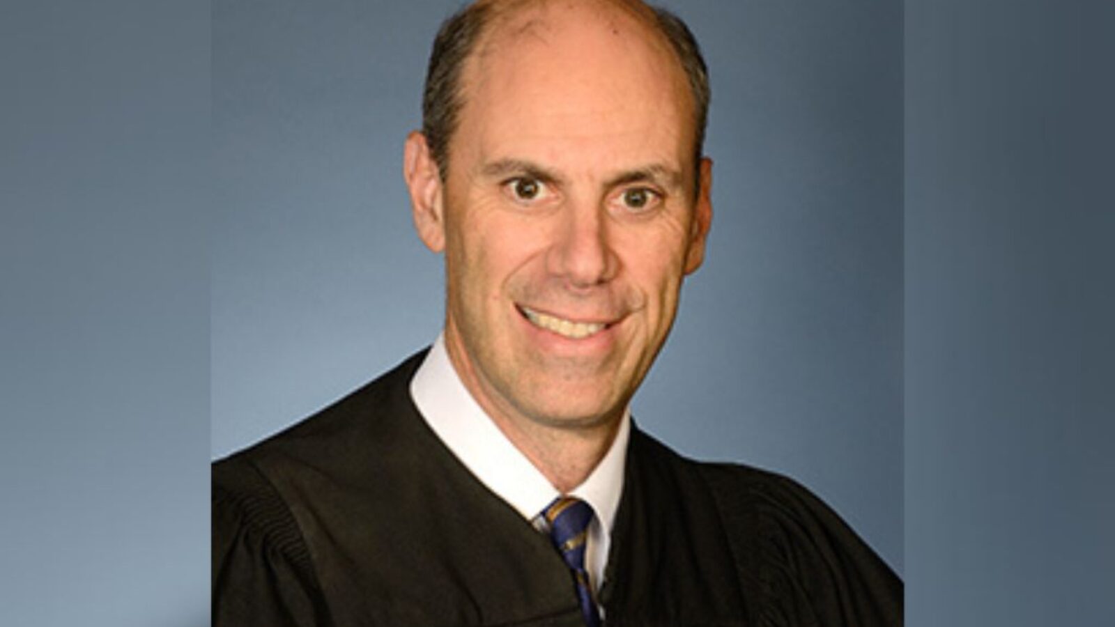 Portrait of a judge in a black robe, smiling against a blue background, representing the judicial profession and authority in law.