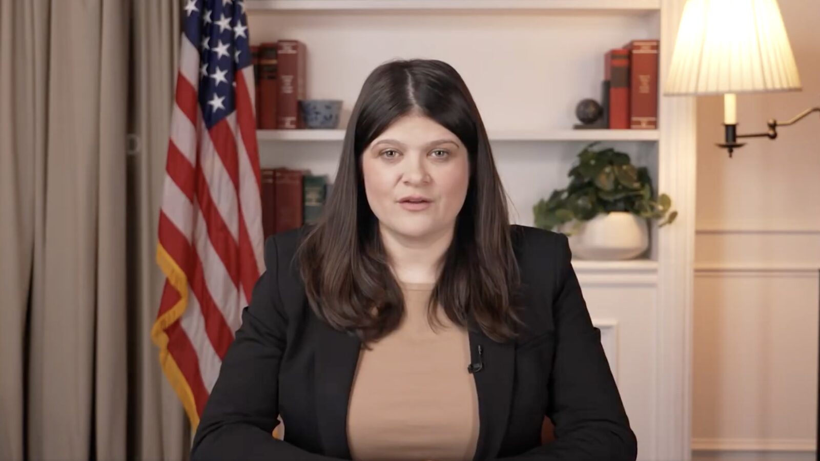 Female speaker in a black blazer sits at a desk with books and a plant, American flag in the background, delivering an address.