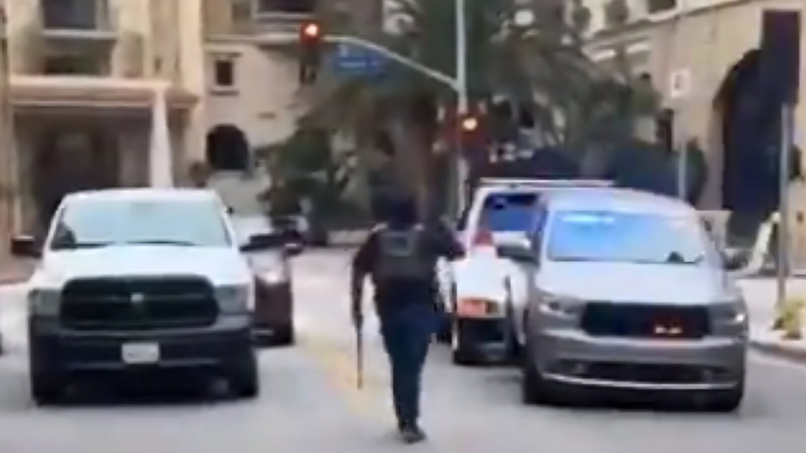 A person in tactical gear walks down a street surrounded by parked vehicles, with palm trees and buildings in the background.