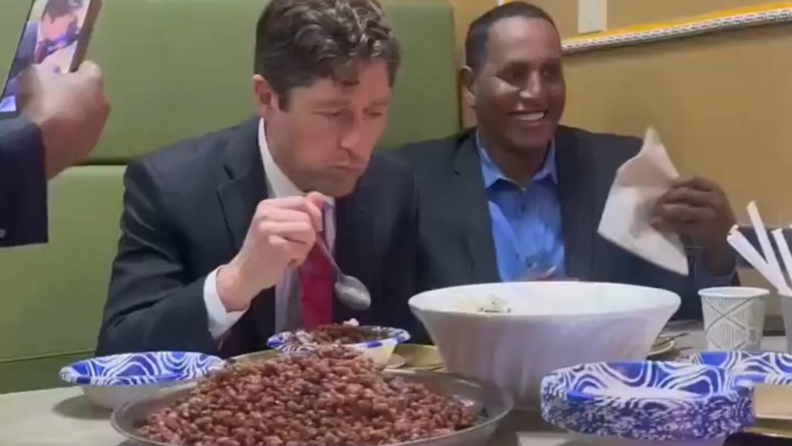 Two men dining at a restaurant, one enjoying a meal of rice and beans, while the other smiles and holds a napkin, capturing a friendly atmosphere.
