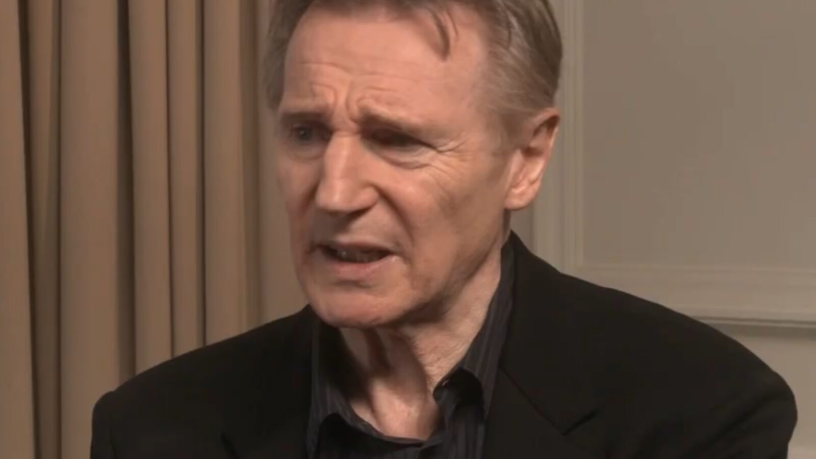 Close-up of a male actor expressing concern during an interview, wearing a black jacket against a neutral background.