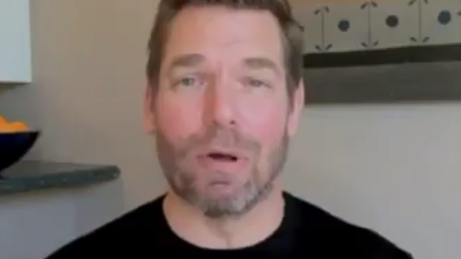 Man with a beard wearing a black shirt speaking in a well-lit room with a bowl of oranges in the background.