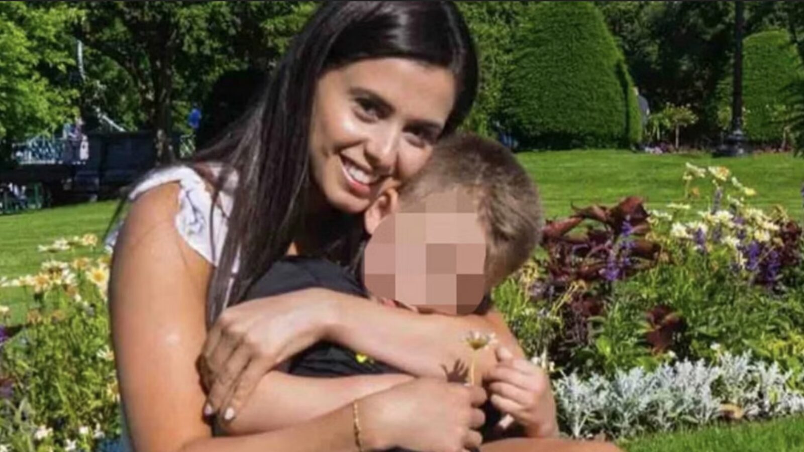 A smiling woman holds a young boy in a park filled with colorful flowers and greenery, showcasing a moment of affection and joy.