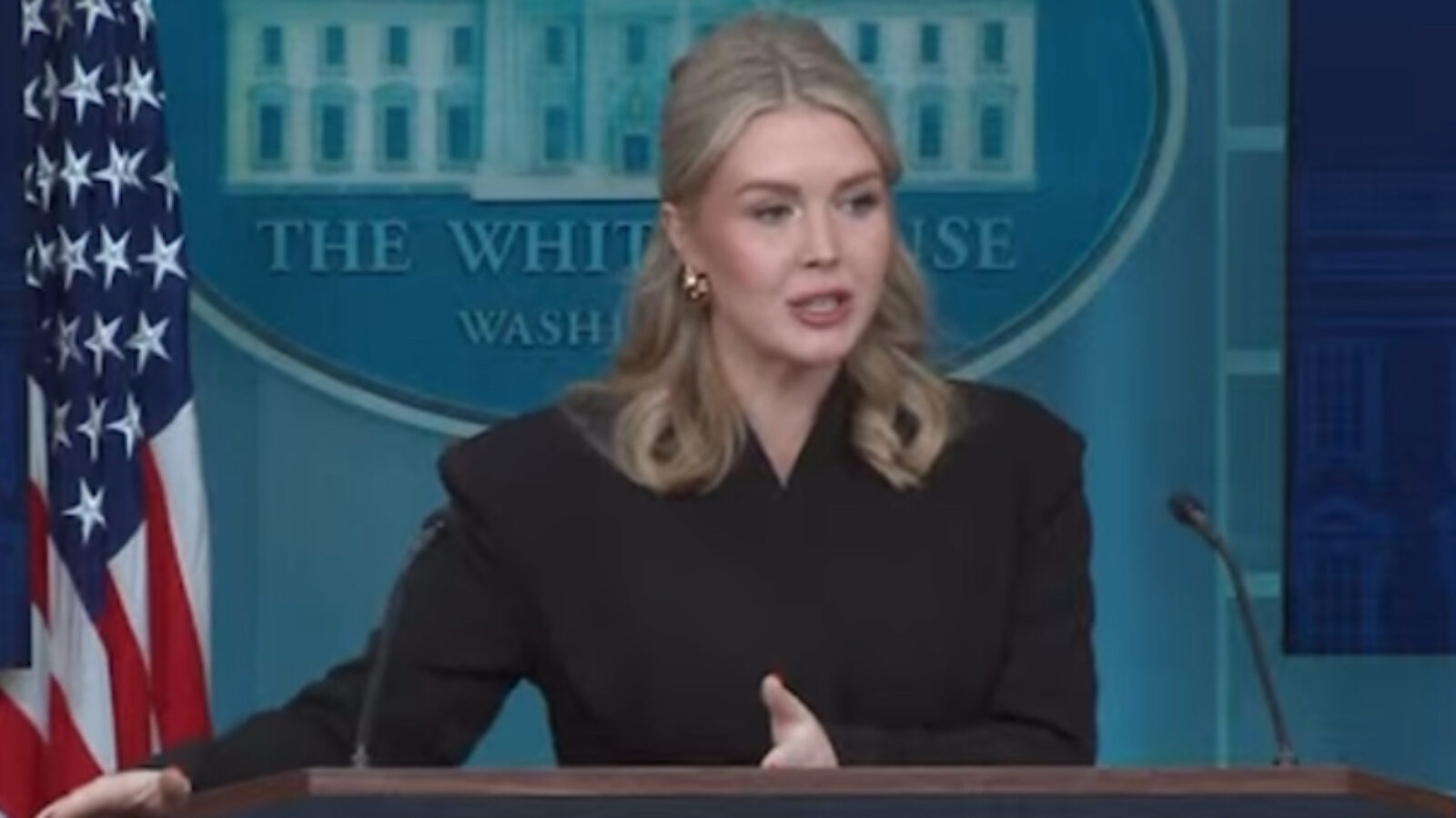 Press secretary delivering a statement at the White House briefing room with the presidential seal and American flags in the background.