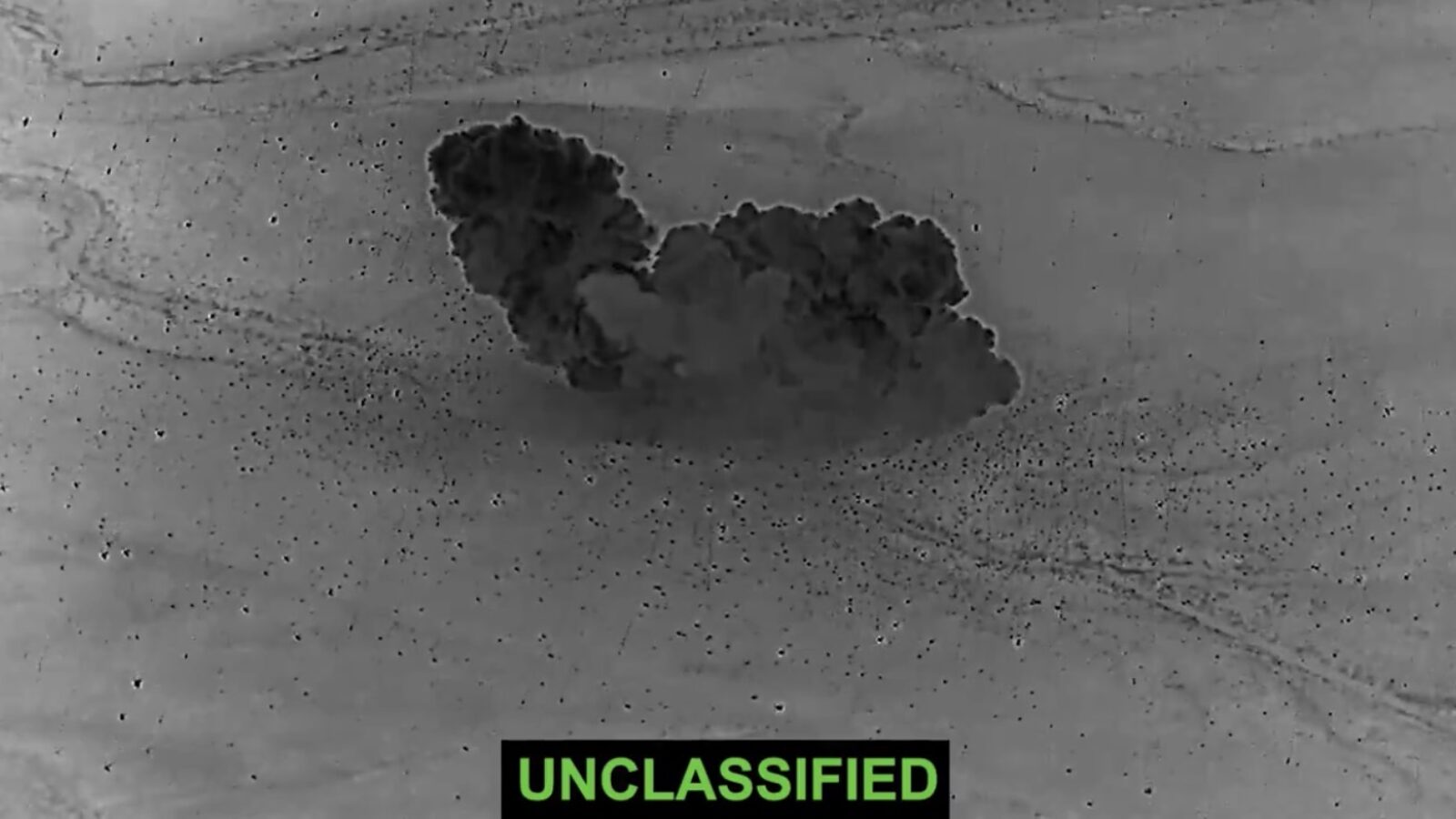 Aerial view of a large explosion producing a dark smoke cloud in a desert landscape, labeled as unclassified.