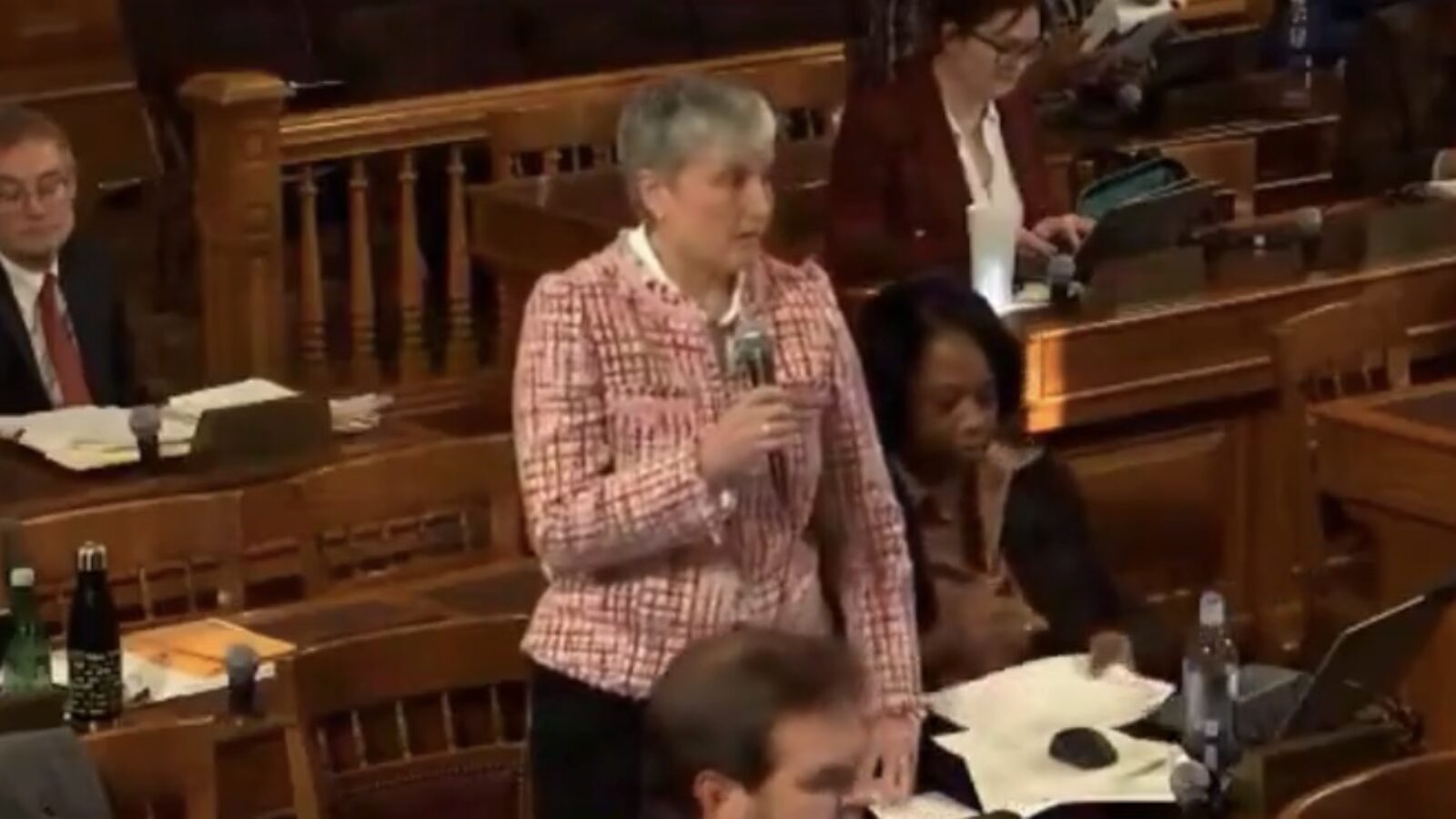 A woman speaks into a microphone in a legislative assembly, with colleagues engaged in discussion and reviewing documents in the background.