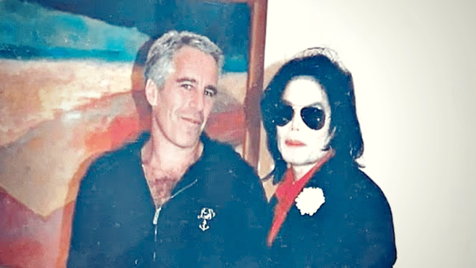 Image of two individuals posing together in front of an abstract painting, one wearing sunglasses and a red shirt, the other in a black jacket.