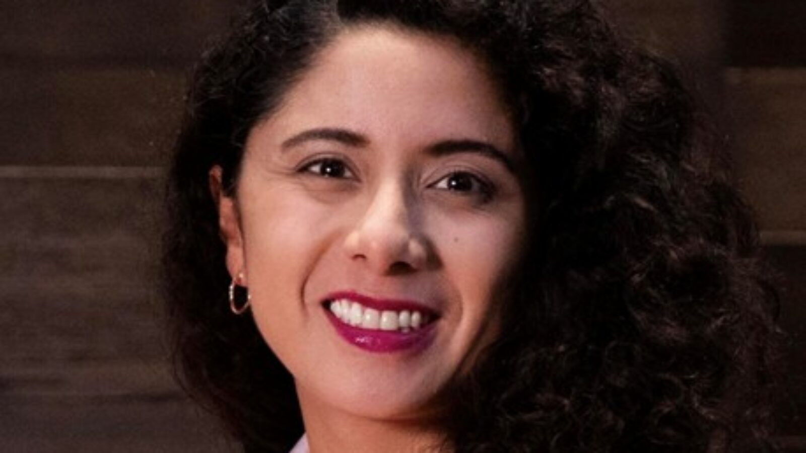 Smiling woman with curly hair wearing a white jacket, posing against a wooden background.