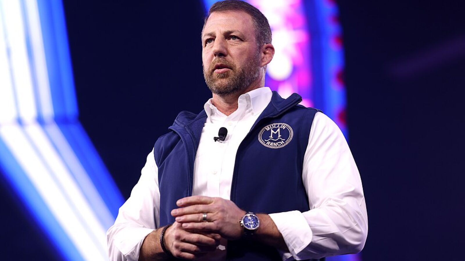Speaker presenting at a conference, wearing a navy vest and white shirt, with a microphone attached, against a colorful background.