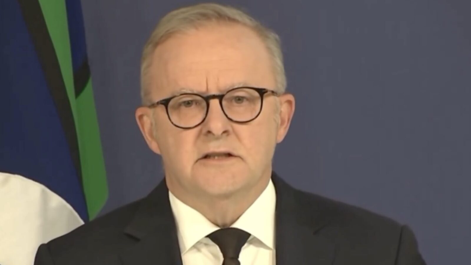 A man in a suit with glasses speaks during a press conference, with a blue and green background featuring flags.