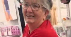 Smiling older woman in a store wearing a red shirt, surrounded by clothing displays and products.