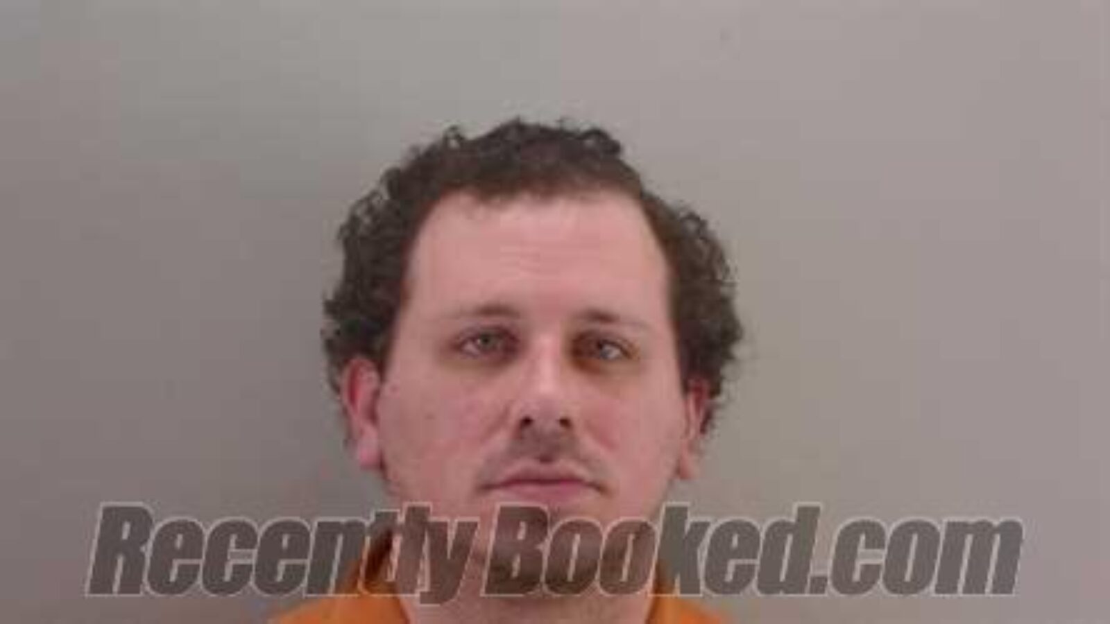 Mugshot of a man with curly hair wearing an orange jumpsuit, captured in a neutral background, typically used for booking records.