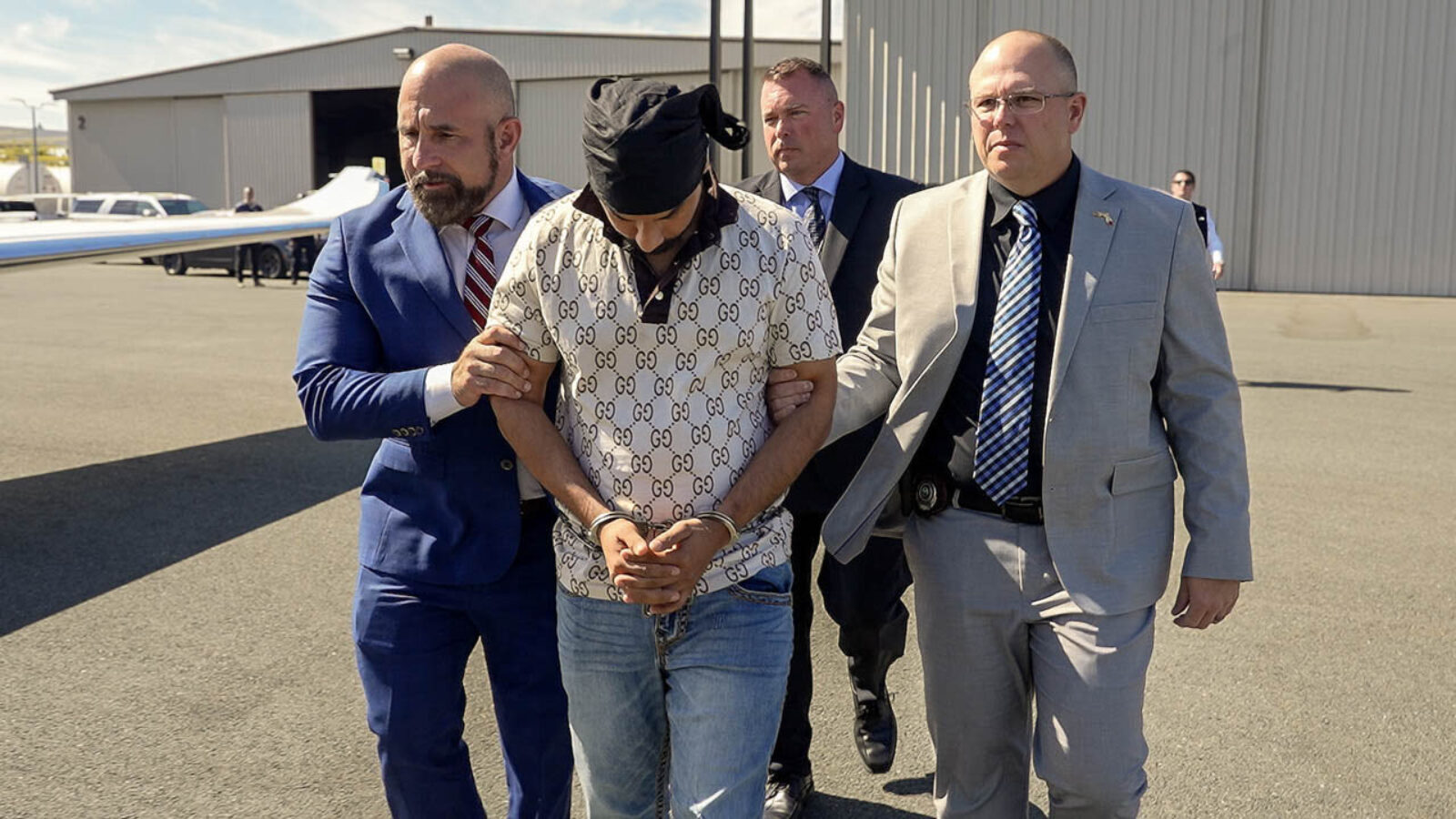 Law enforcement officers escort a handcuffed individual wearing a designer shirt at an airport, highlighting a serious legal situation.