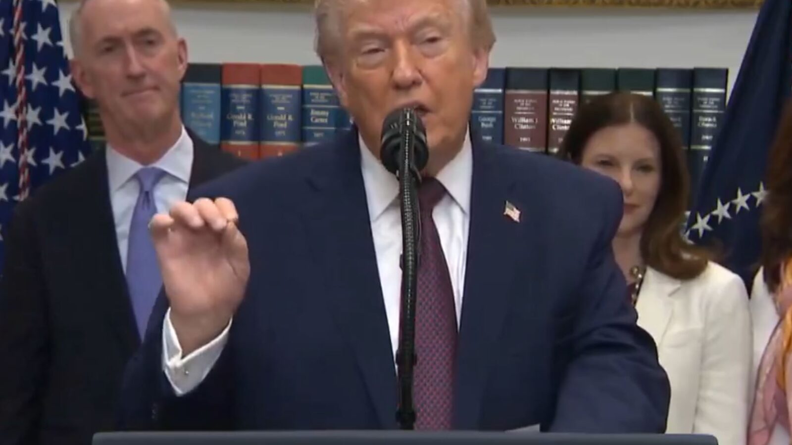 Donald Trump delivers a speech at a podium with the presidential seal, flanked by officials in a formal setting.