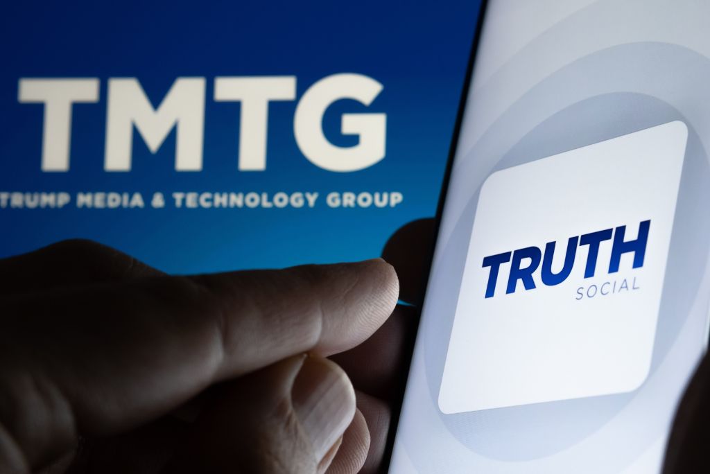 A hand holding a smartphone displays the Truth Social app logo next to the TMTG logo, representing Trump Media & Technology Group.