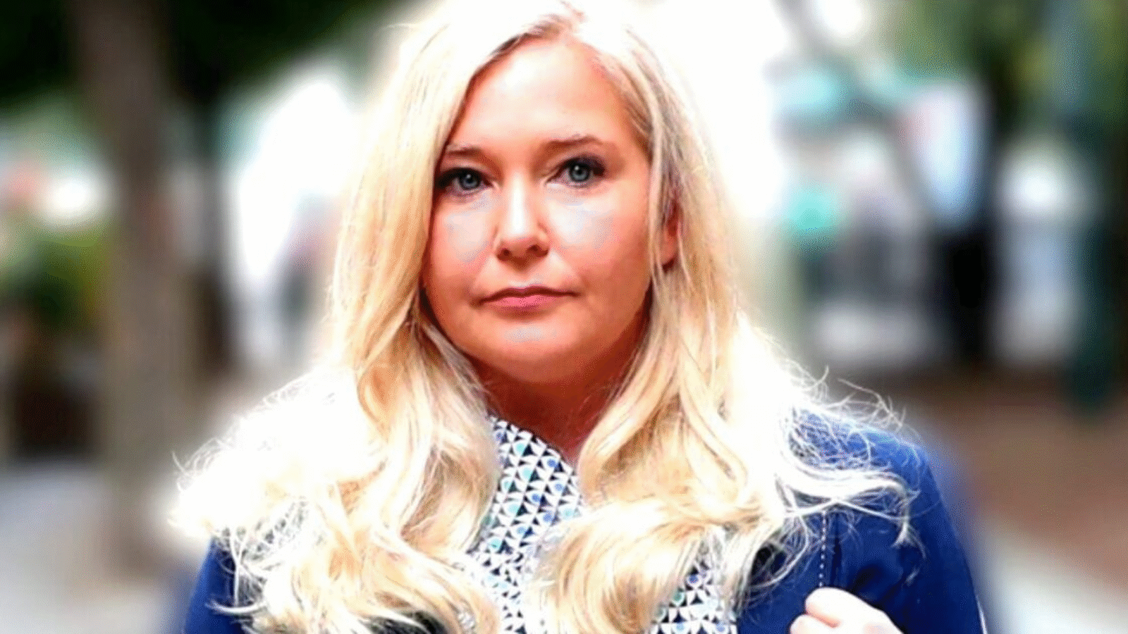 Woman with long blonde hair, wearing a blue blazer and patterned scarf, stands outdoors with a blurred background of trees and people.