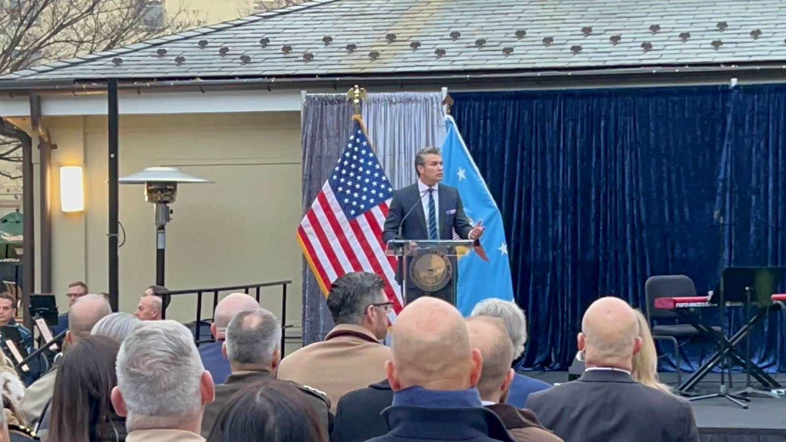 A speaker addresses an audience at an outdoor event featuring American and blue flags, with attendees in formal attire.